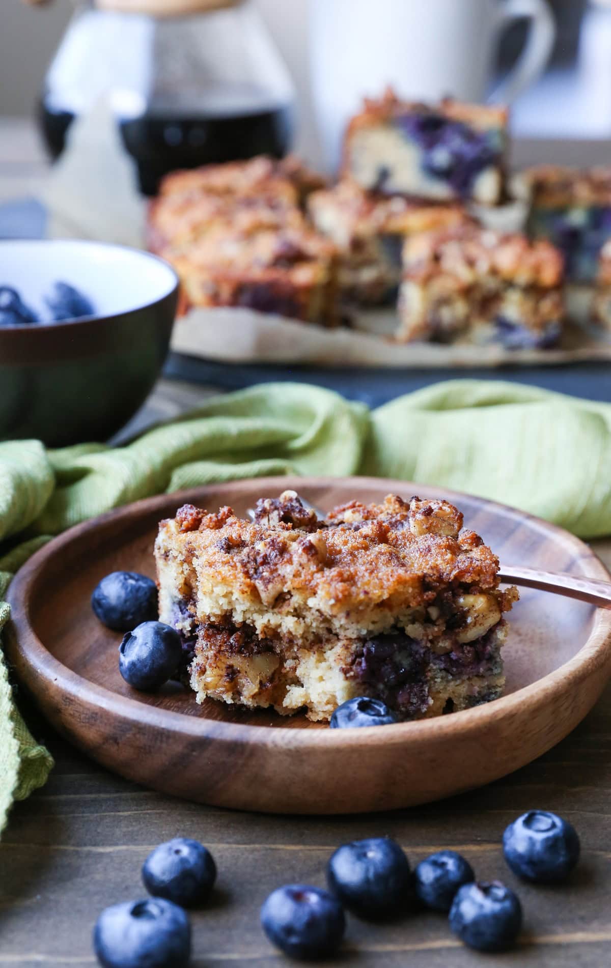 Slice of blueberry coffee cake on a wooden plate with a bite taken out and the rest of the coffee cake in the background with a pour over coffee vessel.