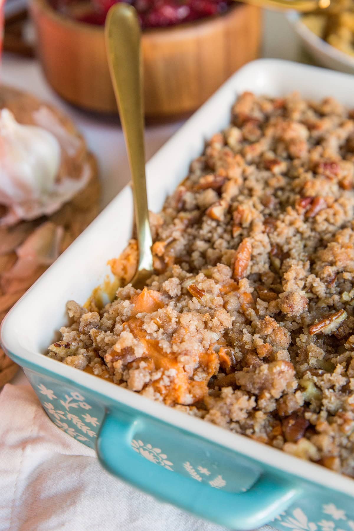 Healthy Paleo Sweet Potato Casserole in a blue casserole dish with a golden spoon scooping some of the casserole out. More Thanksgiving side dishes in the background.
