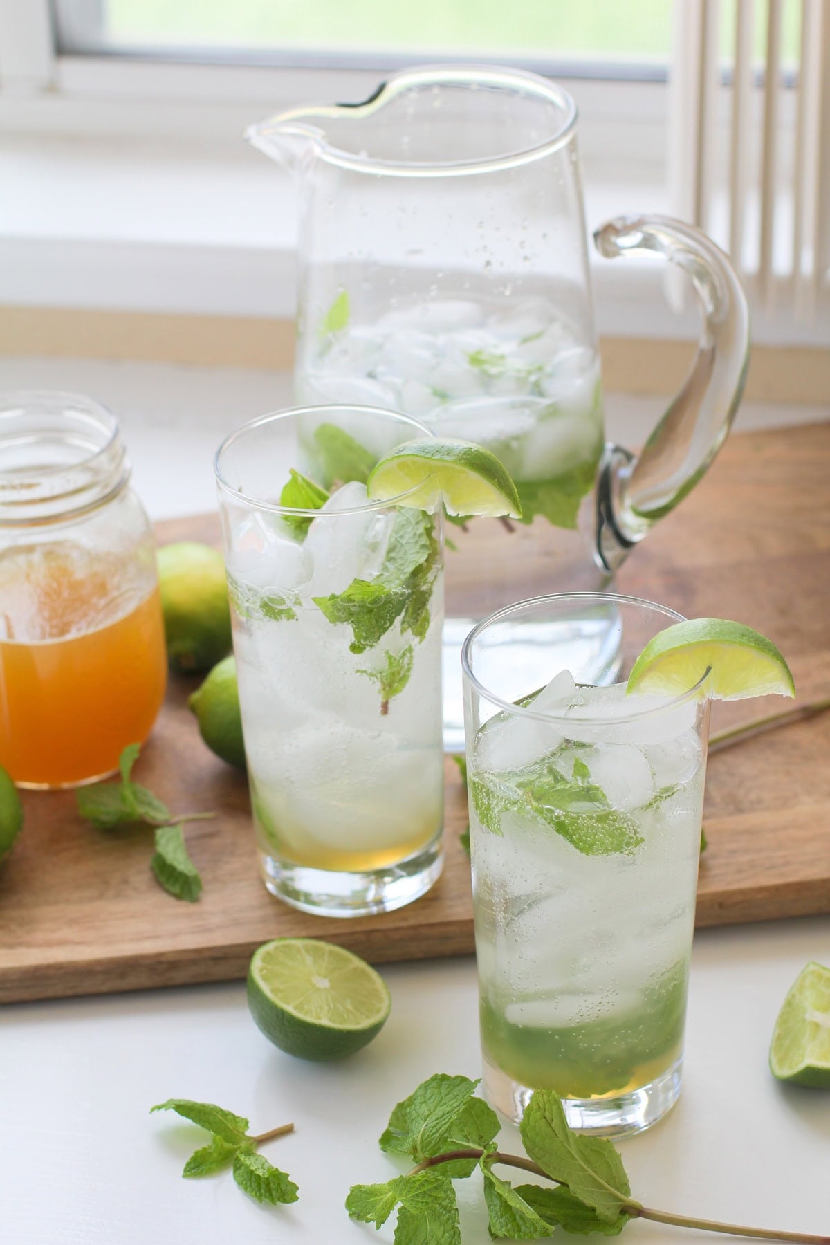 Two glasses of mint mojitos with a pitcher of mojitos in the background and fresh mint leaves to the side.