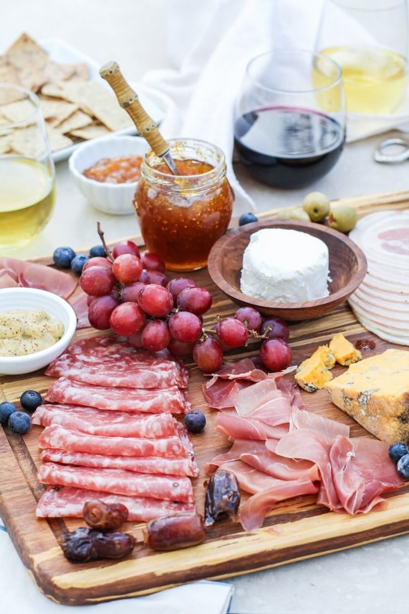 how to make a charcuterie board with meat, cheese, and wine. Large wooden board full of meats, cheeses, spreads, and fruit to create delicious bites of food.