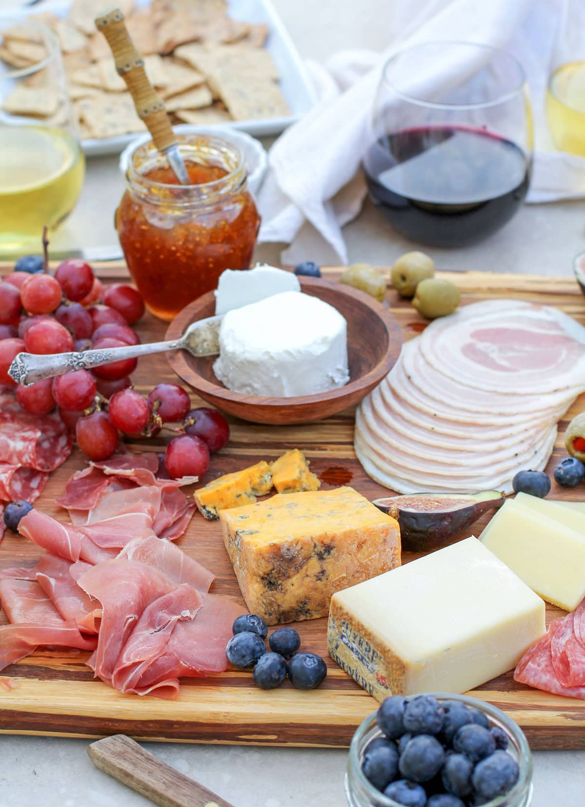 Large meat and cheese board sitting on a table top with a variety of sliced meats, cheeses, fresh fruit, spreads, and glasses of wine for an amazing appetizer experience