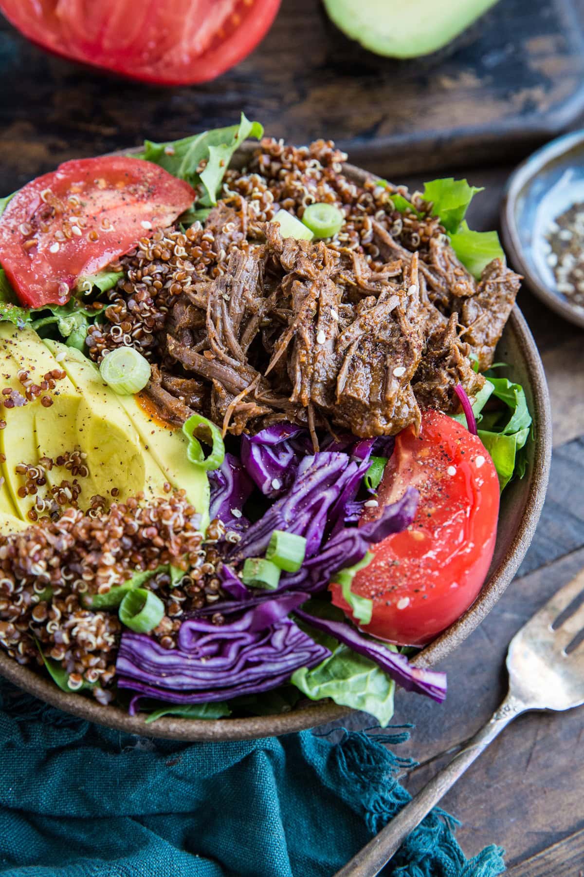 Big bowl of greens, quinoa, barbacoa beef, avocado, tomatoes with a fork to the side and a blue napkin, ready to serve.