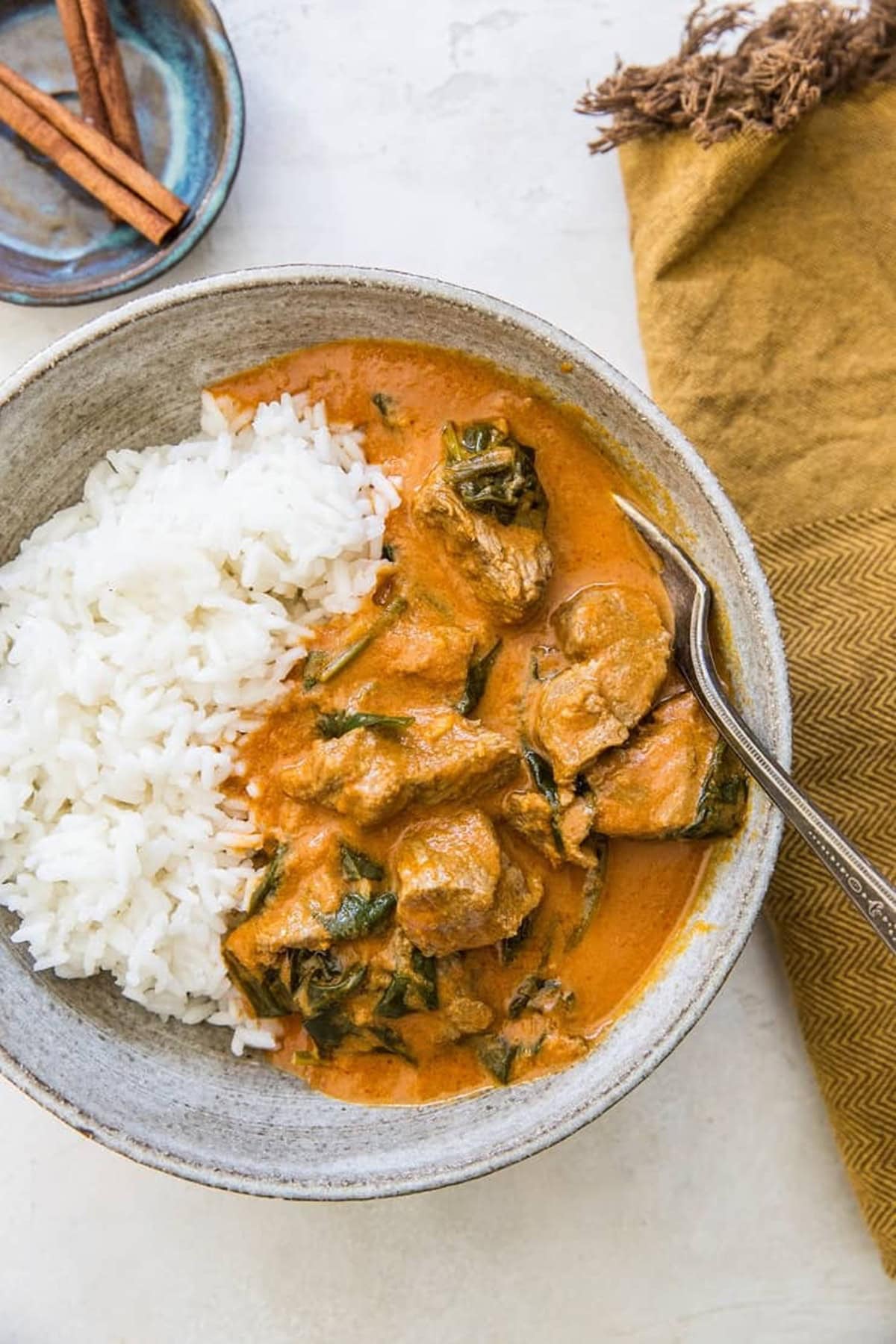 Large bowl of lamb curry with white rice with a golden napkin to the side and a bowl of cinnamon sticks.