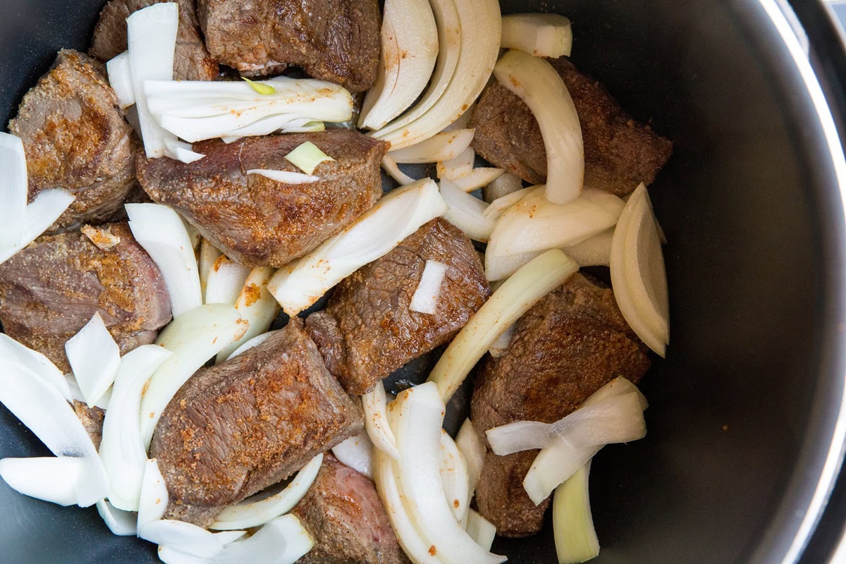 Chunks of browned beef and sliced onions are arranged in a black pot, ready to be cooked together for a savory Instant Pot Pot Roast.