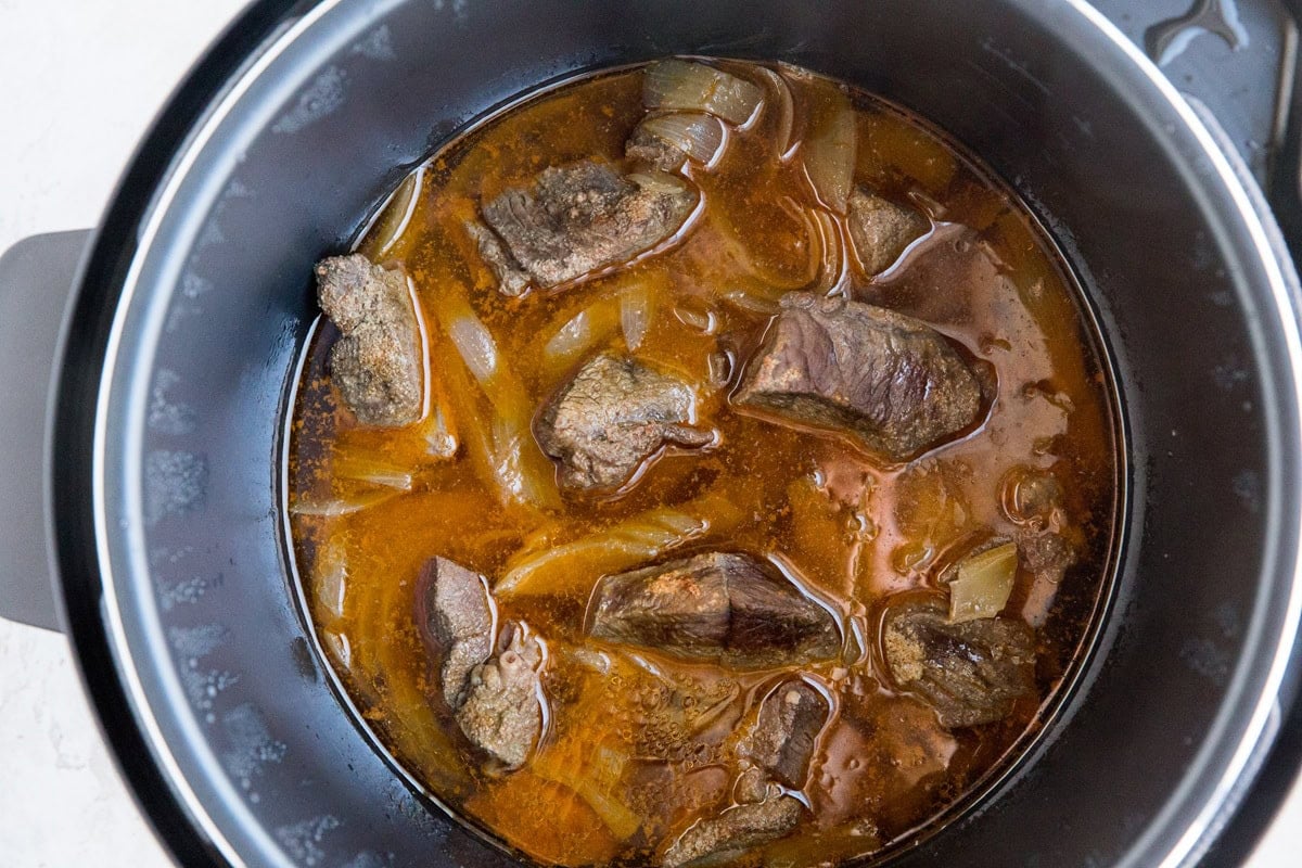 Chunks of beef and chopped onions simmer in a rich, brown broth inside a black Instant Pot Pot Roast slow cooker or pressure cooker.