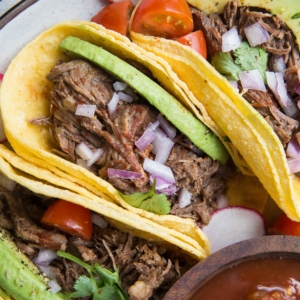 Three shredded beef tacos on a plate with tomato salsa, avocado, cilantro, and red onion. Ready to eat.