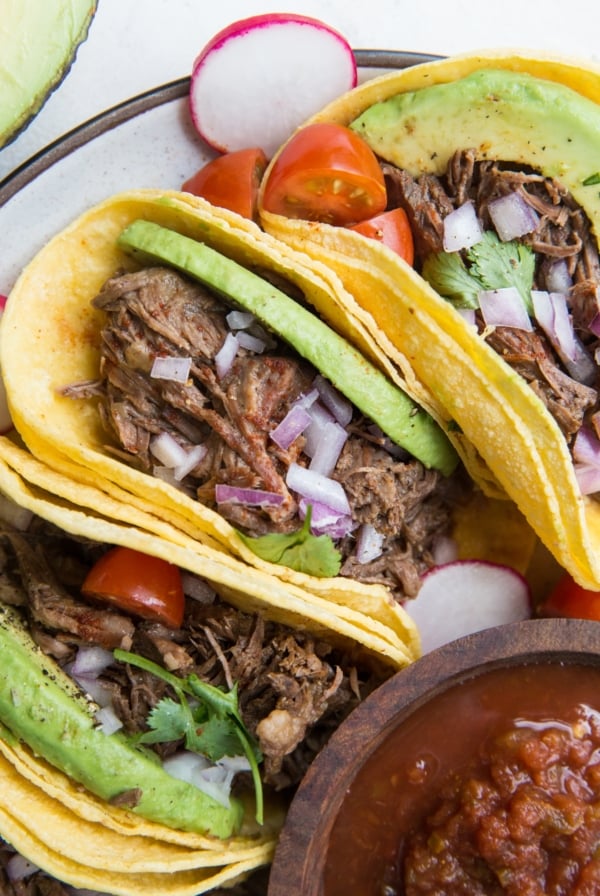 Three shredded beef tacos on a plate with tomato salsa, avocado, cilantro, and red onion. Ready to eat.