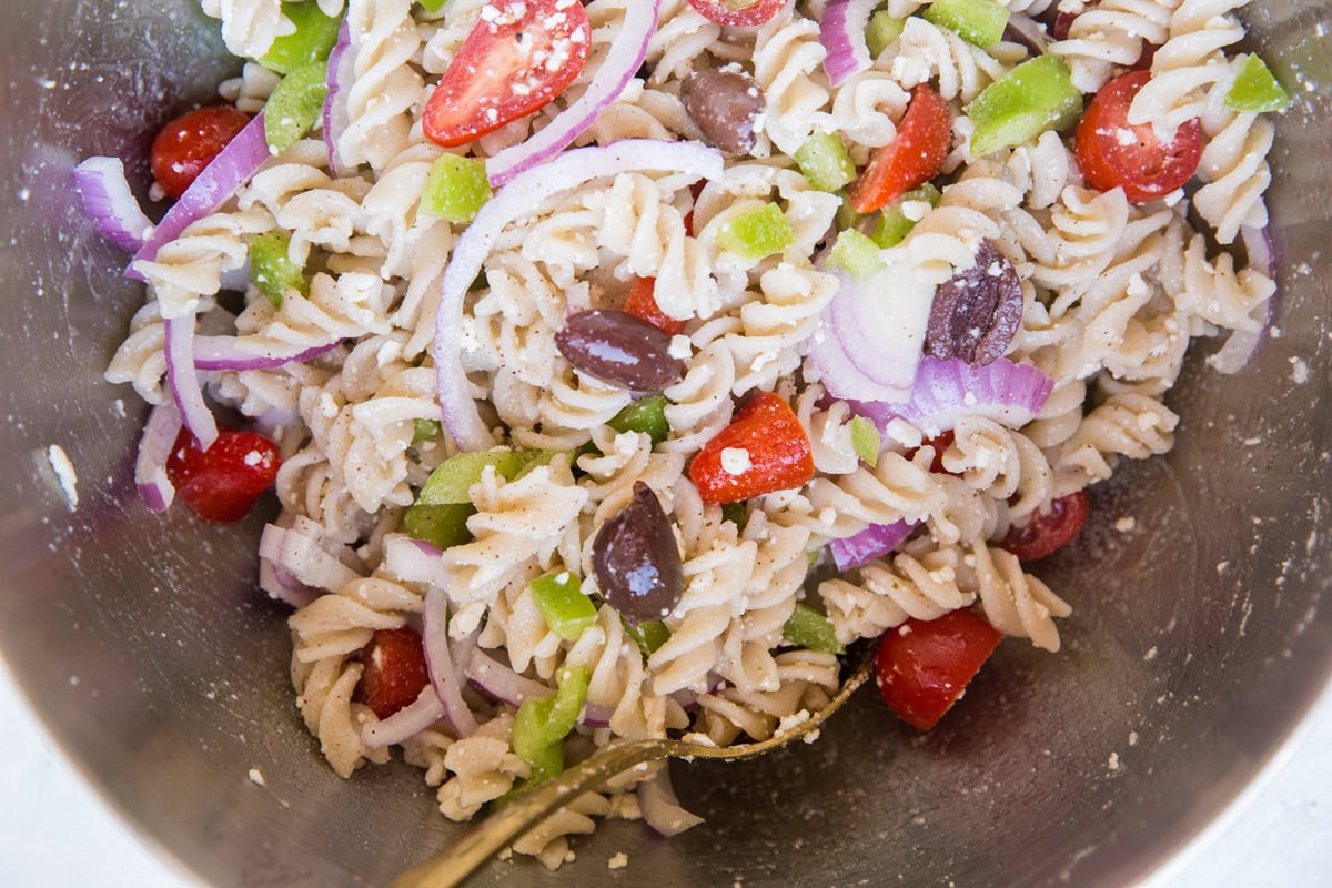 Italian pasta salad ingredients tossed together in a mixing bowl. Cooked pasta, kalamata olives, bell peppers, red onion, cherry tomatoes and feta cheese with Italian dressing.