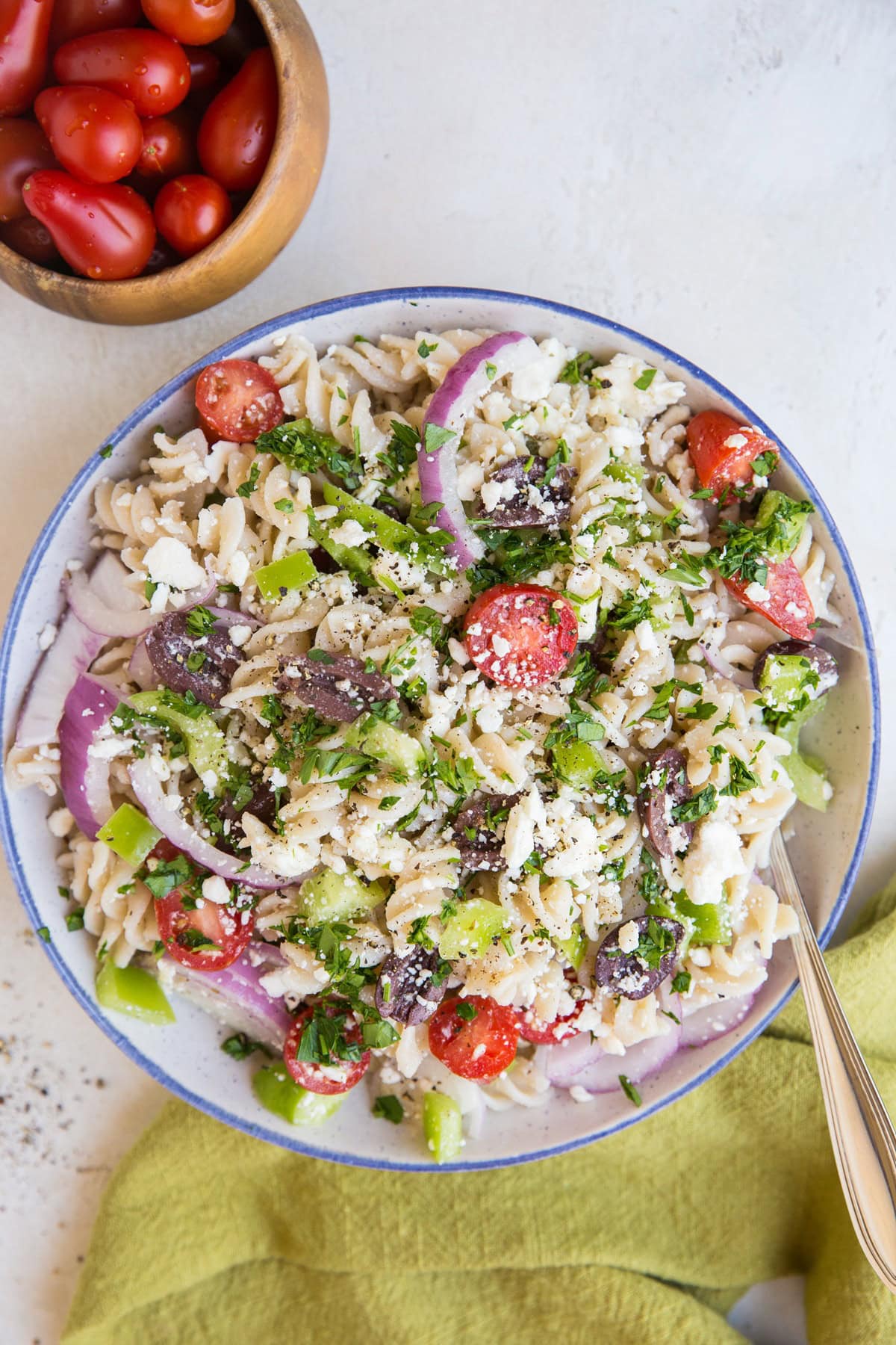 Large bowl full of pasta salad with a serving spoon inside. A green napkin and a bowl of fresh cherry tomatoes to the side.