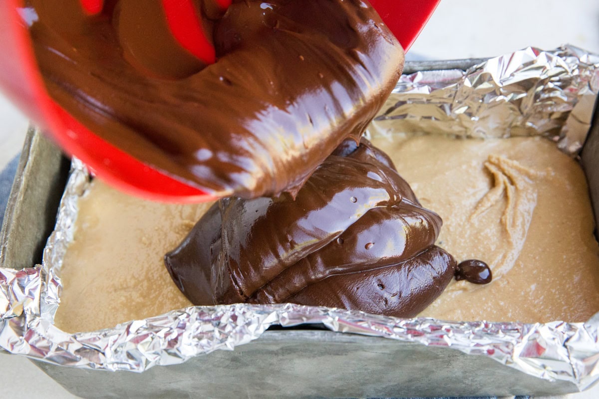 Pouring chocolate ganache into a loaf pan with peanut butter mixture to create a chocolate layer.
