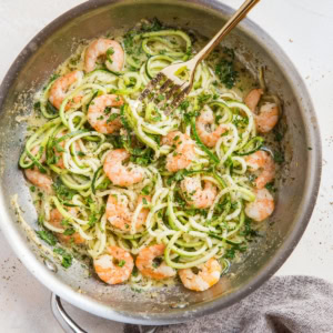 Stainless steel skillet full of zucchini noodles and shrimp with a fork twirling some of the noodles and a grey napkin to the side.