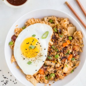 Large white bowl full of kimchi fried rice with a fried egg on top and sprinkles of sesame seeds to the side.