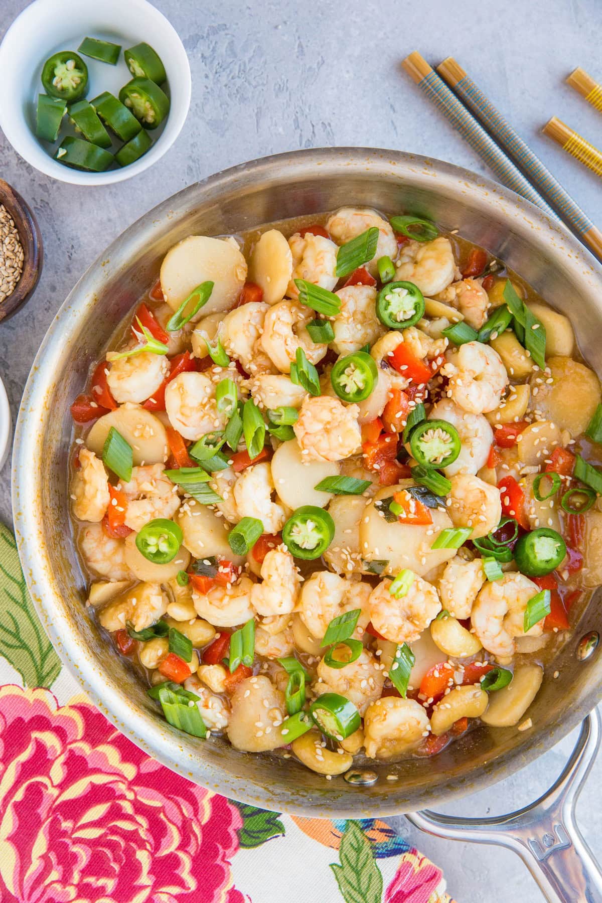 Skillet full of kung pao shrimp with a bright napkin to the side, chop sticks, and a bowl of green chiles