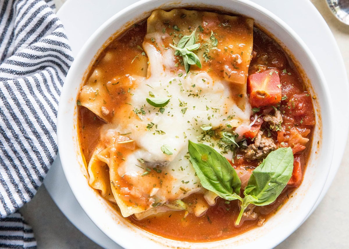 Lasagna Soup in a white bowl with melted cheese on top and a fresh basil leaf. Striped napkin to the side.