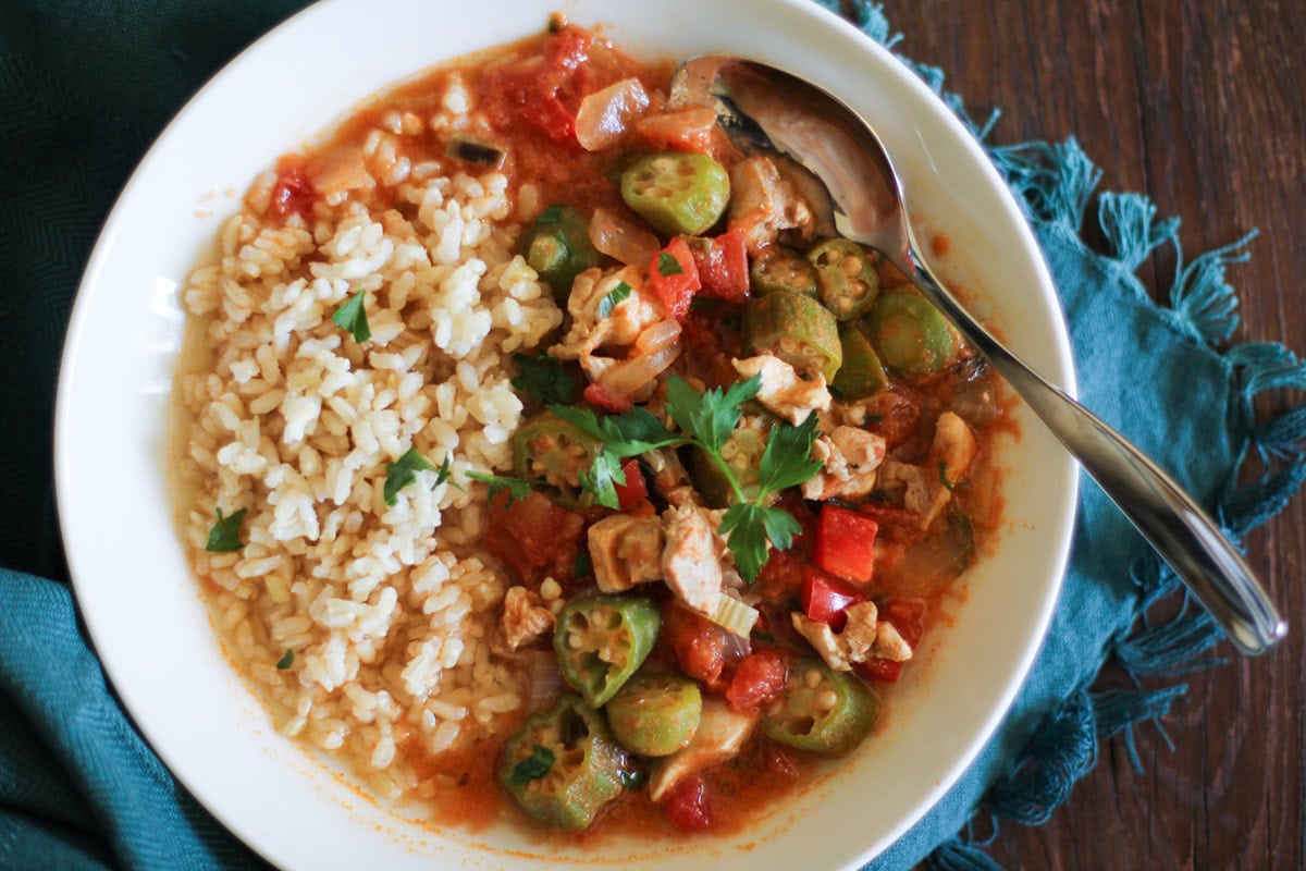 Louisiana-Style Chicken Soup in a large white bowl with brown rice to the side and a silver spoon and a blue napkin, ready to eat.