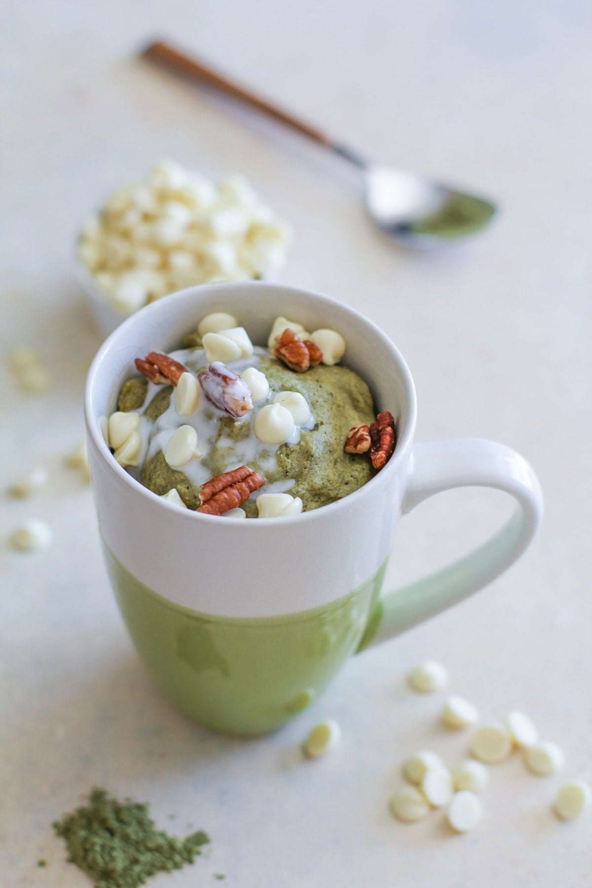 Gluten-Free and Naturally Sweetened Matcha Mug Cake in a green and white mug with white chocolate chips and matcha powder sprinkled around.