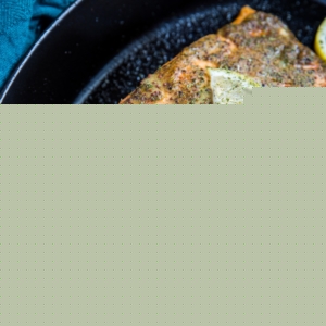 Large salmon filet in a baking dish with a coat of mustard and seasonings on top for a baked mustard salmon recipe. A fork taking bites at the fish to show how flaky and moist it is. A slice of lemon on top of the fish.