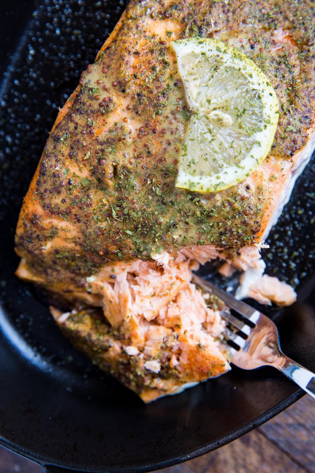 Mustard Baked salmon with lemon in a baking dish with a slice of lemon on top. A fork taking a bite out of the fish to reveal the flaky nature of it.