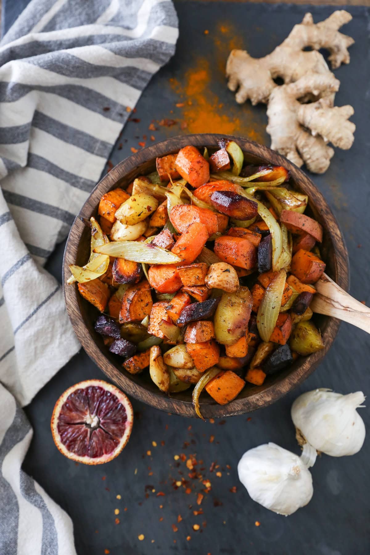 Wooden bowl full of roasted root vegetables with fresh garlic, a blood orange, and ginger to the side. Ready to serve.