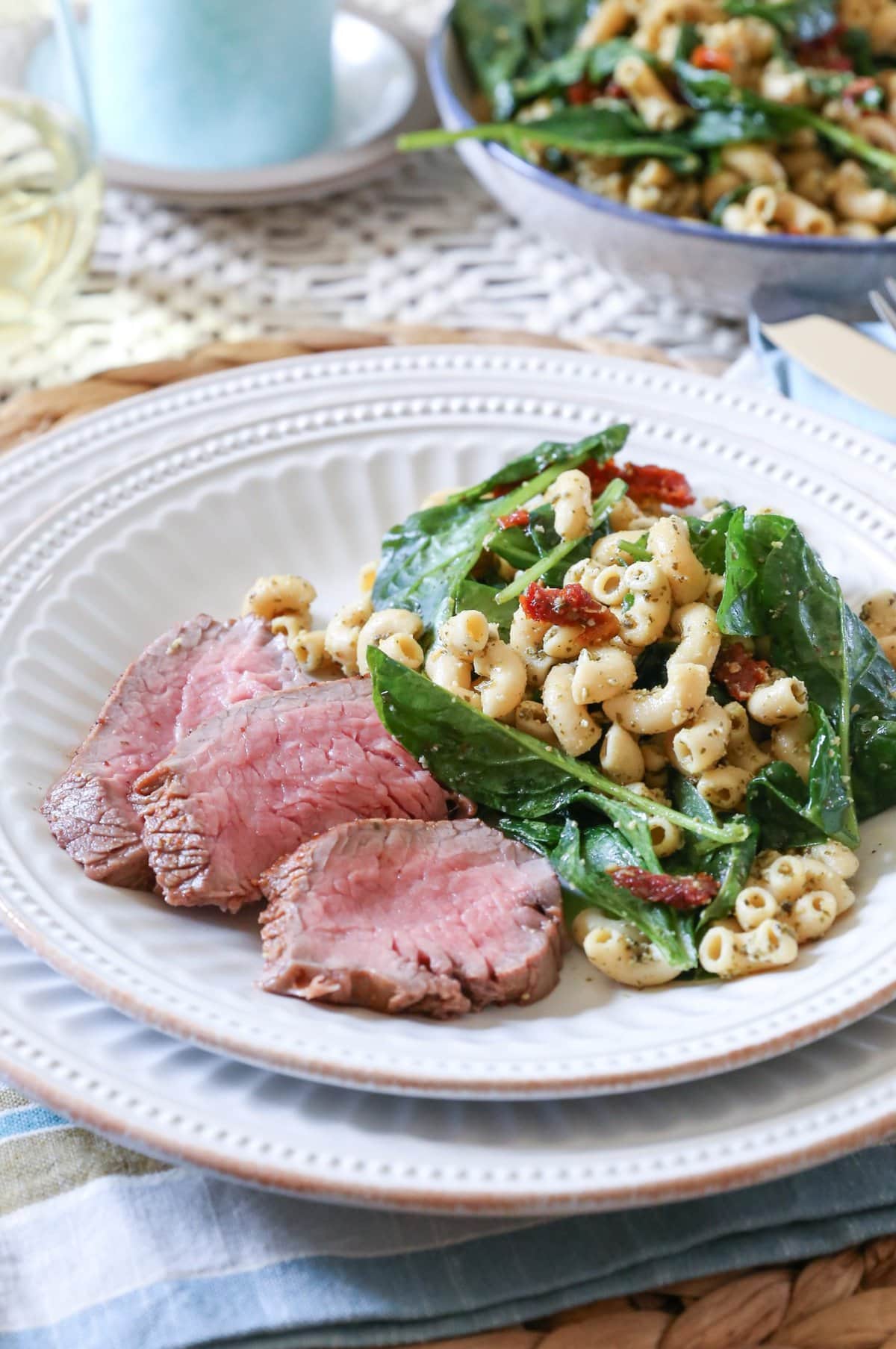 Slices of oven baked tri tip on a white plate with macaroni salad. A place mat and napkin underneath the plate and a glass of wine in the background.
