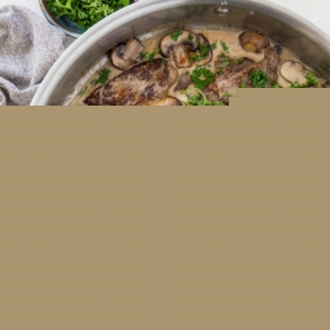 A stainless steel skillet filled with creamy paleo beef stroganoff, featuring sliced beef, mushrooms, onions, and a garnish of chopped parsley. A small bowl of fresh parsley and a gray cloth sit beside the skillet.