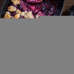 Paleo Blackberry Cobbler in two white bowls, one with a scoop of vanilla ice cream on top. The rest of the cobbler in a baking dish and fresh blackberries and spoons to the side.