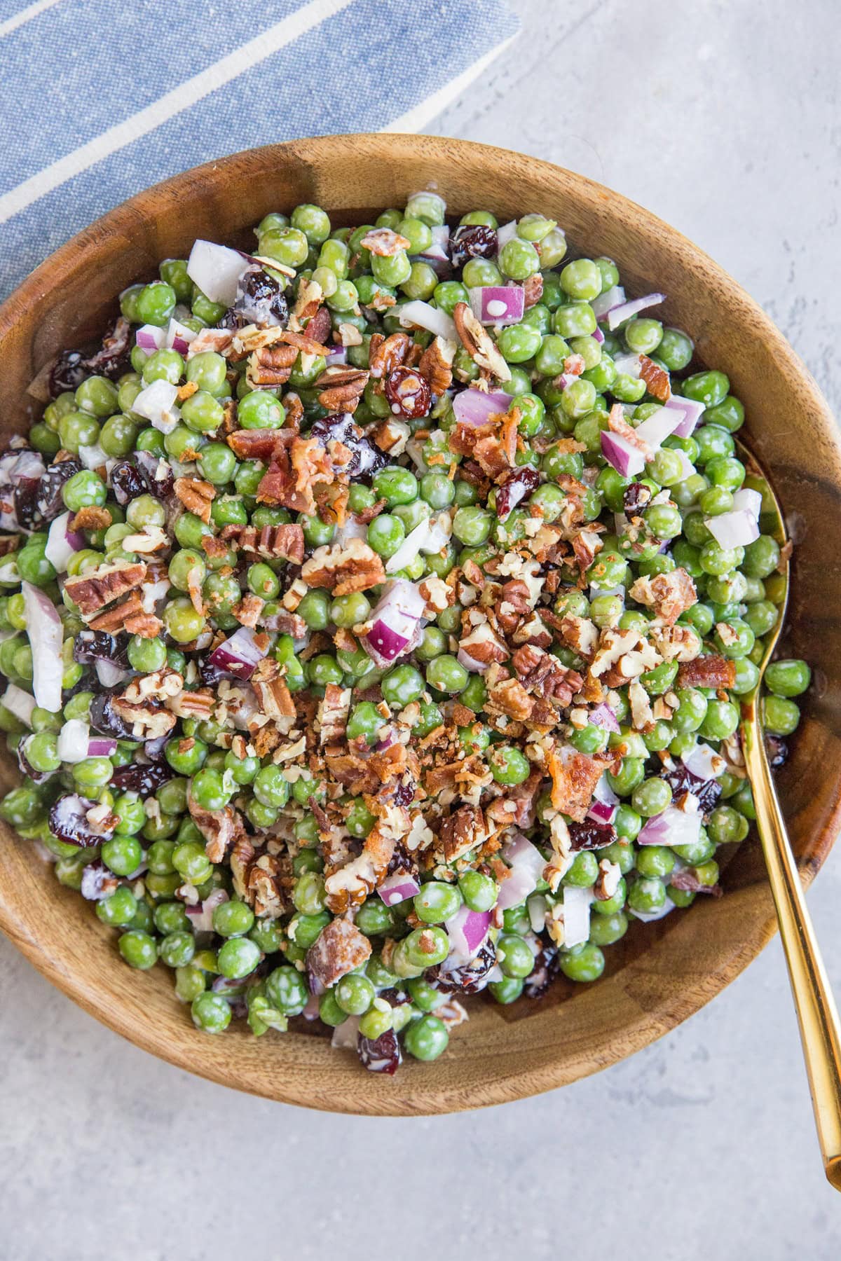 Mayo-Free Pea Salad with Bacon, dried cranberries, pecans, and red onion in a wooden bowl with a golden spoon and a blue striped napkin to the side. Ready to serve.