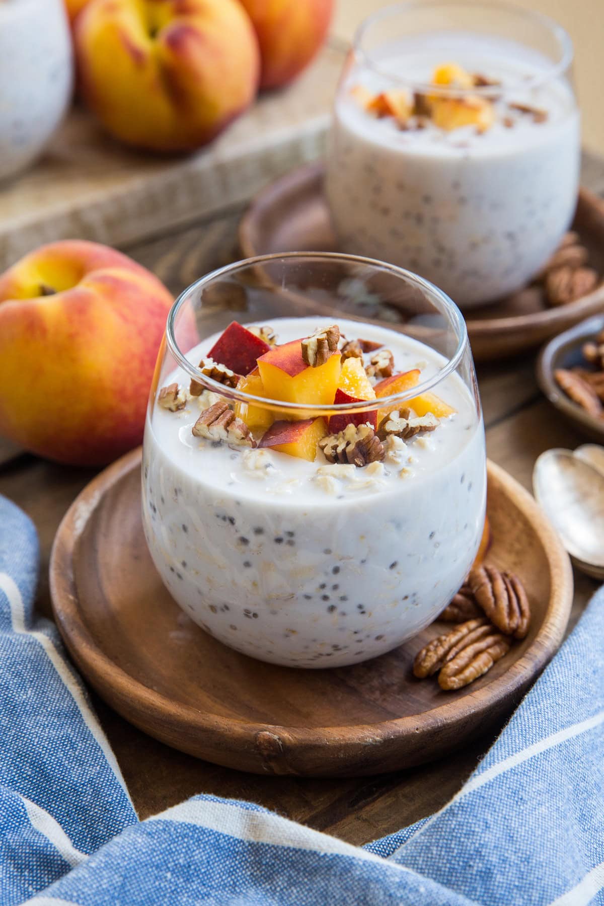 Two glasses of peaches and cream overnight oats with pecans and fresh chopped peaches on top with spoons and a blue striped napkin.
