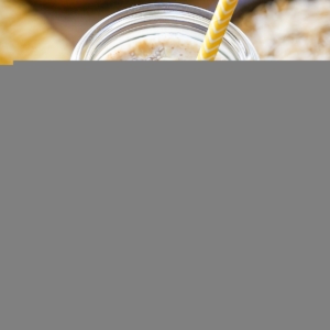Banana Bread Oat Protein Smoothie in a glass with a yellow striped straw, ready to drink. Peanut butter, oats, and bananas in the background.
