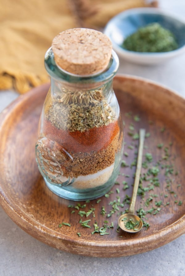 Jar full of seasonings with a cork on top and a small spoon to the side with dried parsley around the jar of seasonings and a small bowl of dried parsley in the background.
