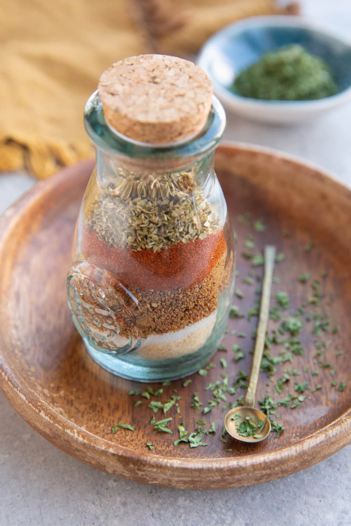 Jar full of seasonings with a cork on top and a small spoon to the side with dried parsley around the jar of seasonings and a small bowl of dried parsley in the background.