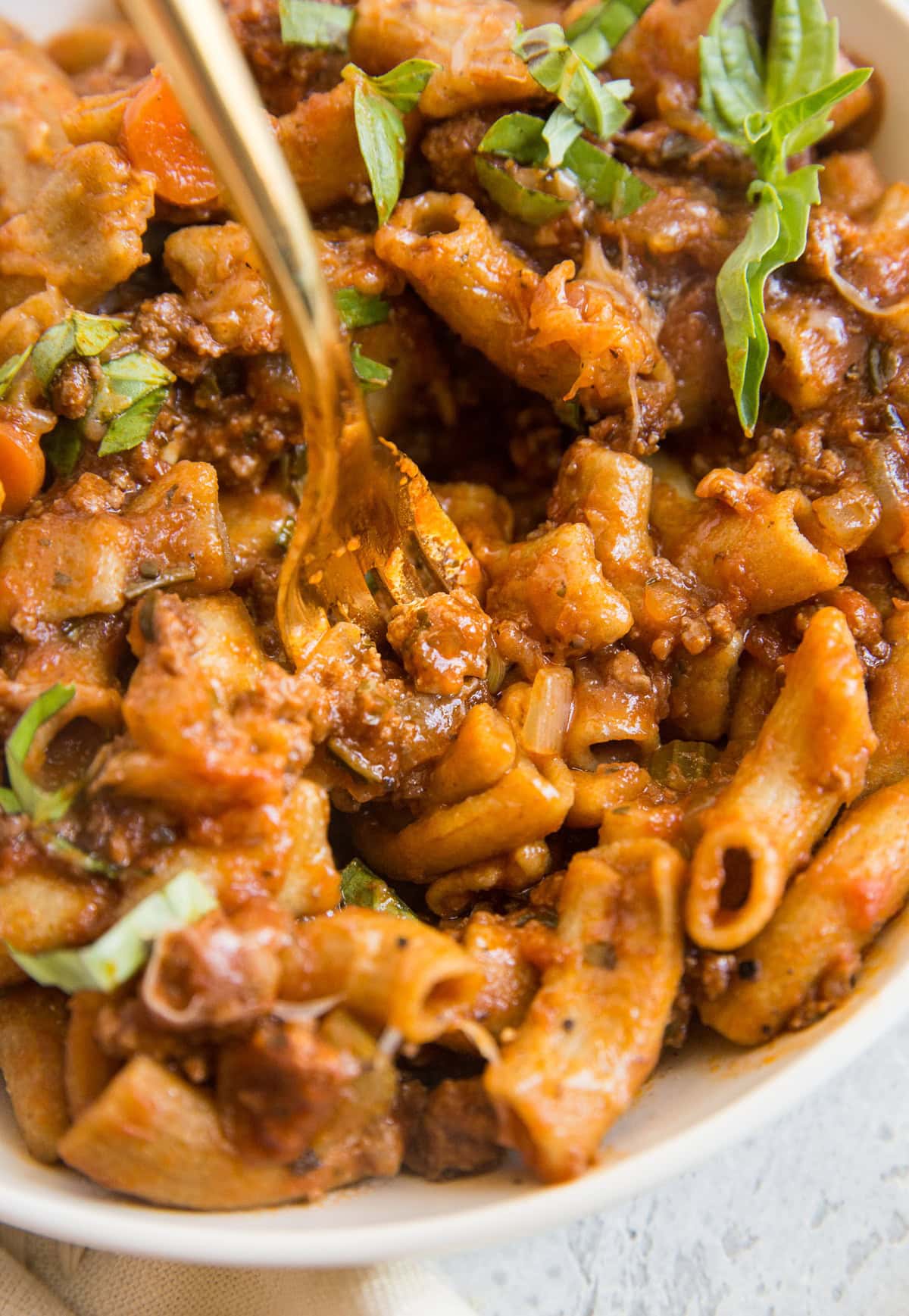 Rigatoni Bolognese in a large white bowl with a golden fork taking a bite.