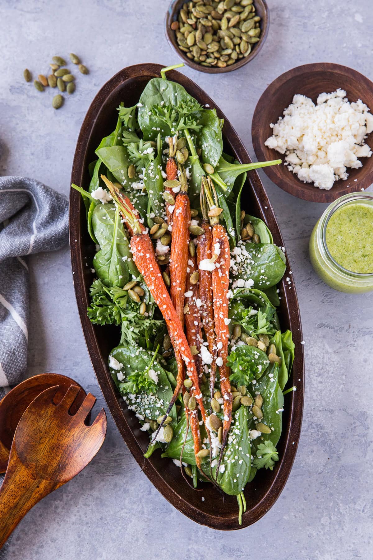 large wooden serving bowl full of spinach and carrot salad. Salad tongs to the side and a bowl of feta cheese and a jar of dressing.