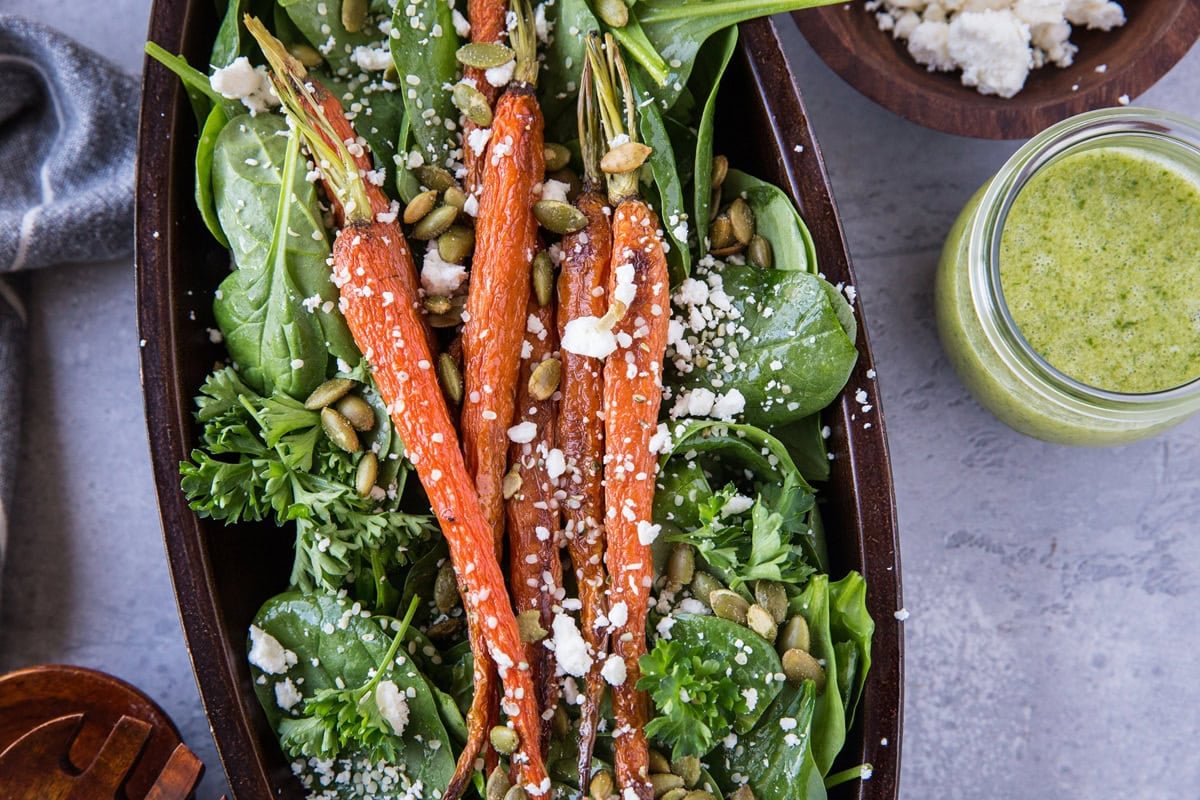 large wooden bowl full of spinach salad with roasted carrots on top, sprinkled with pumpkin seeds and feta cheese. Ready to serve.