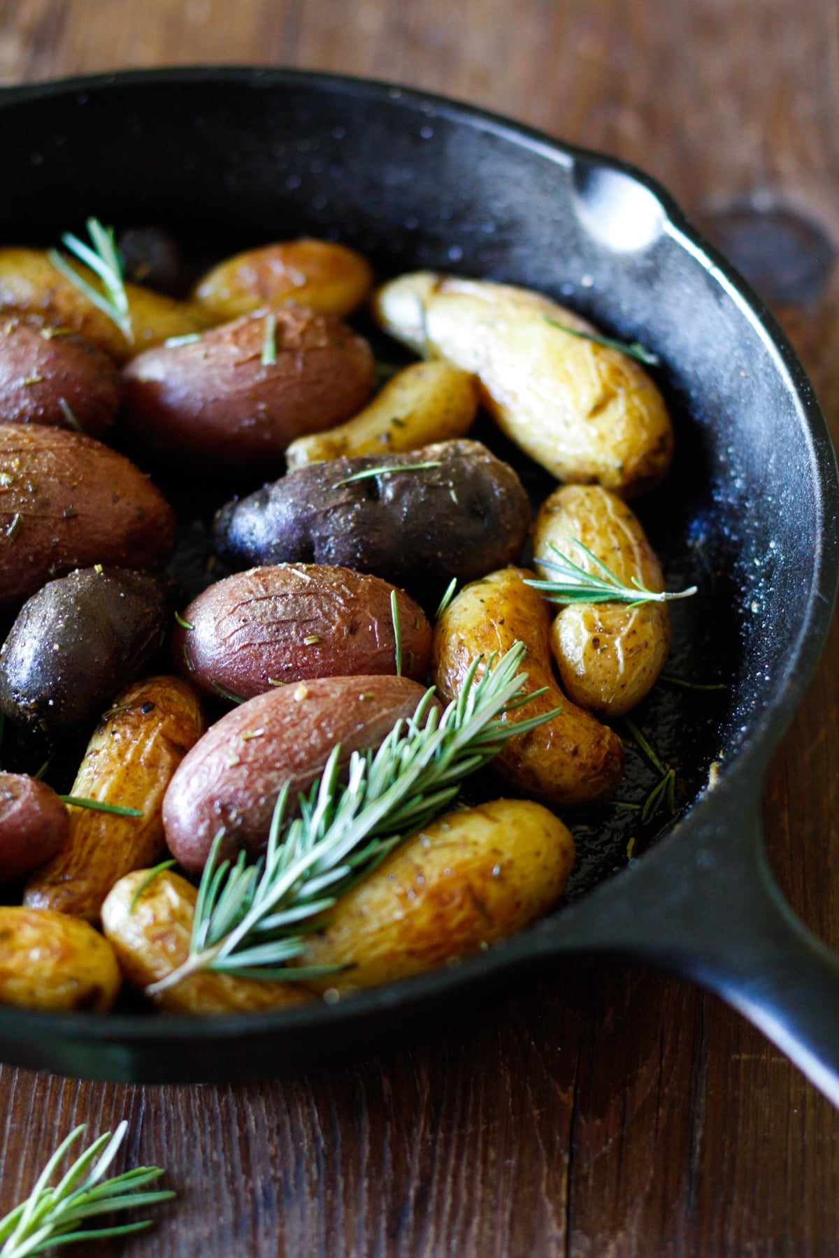A black cast iron skillet filled with roasted fingerling potatoes in various colors, garnished with fresh sprigs of rosemary, sits on a rustic wooden surface. The roasted fingerling potatoes add vibrant appeal to the scene.