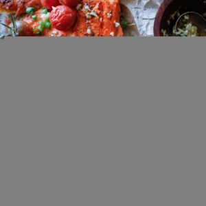 Long salmon filet on a black cutting board topped with cooked tomatoes, garlic, and chopped green onions, and cut into individual filets. Ready to serve.