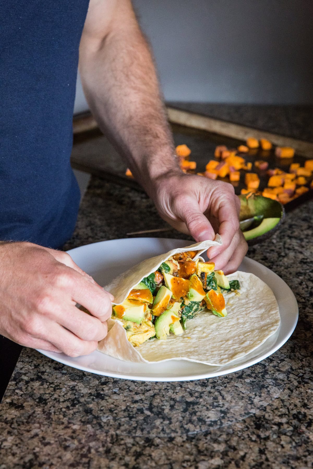 Man folding a tortilla over scrambled eggs, vegetables, and avocado with hot sauce. Folding in the edges of the tortilla first.