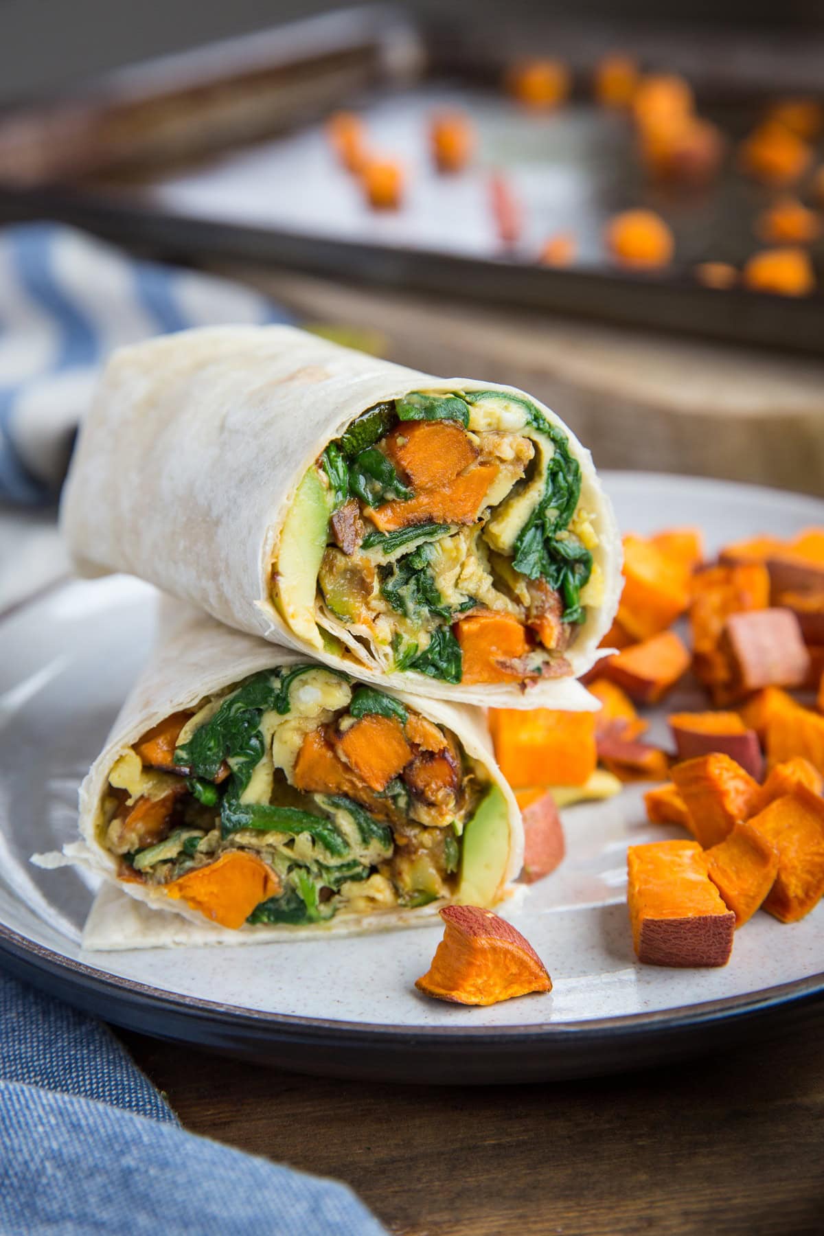 Roasted vegetable breakfast burrito cut in half on a plate so you can see the inside. Roasted sweet potatoes to the side of the breakfast burrito.