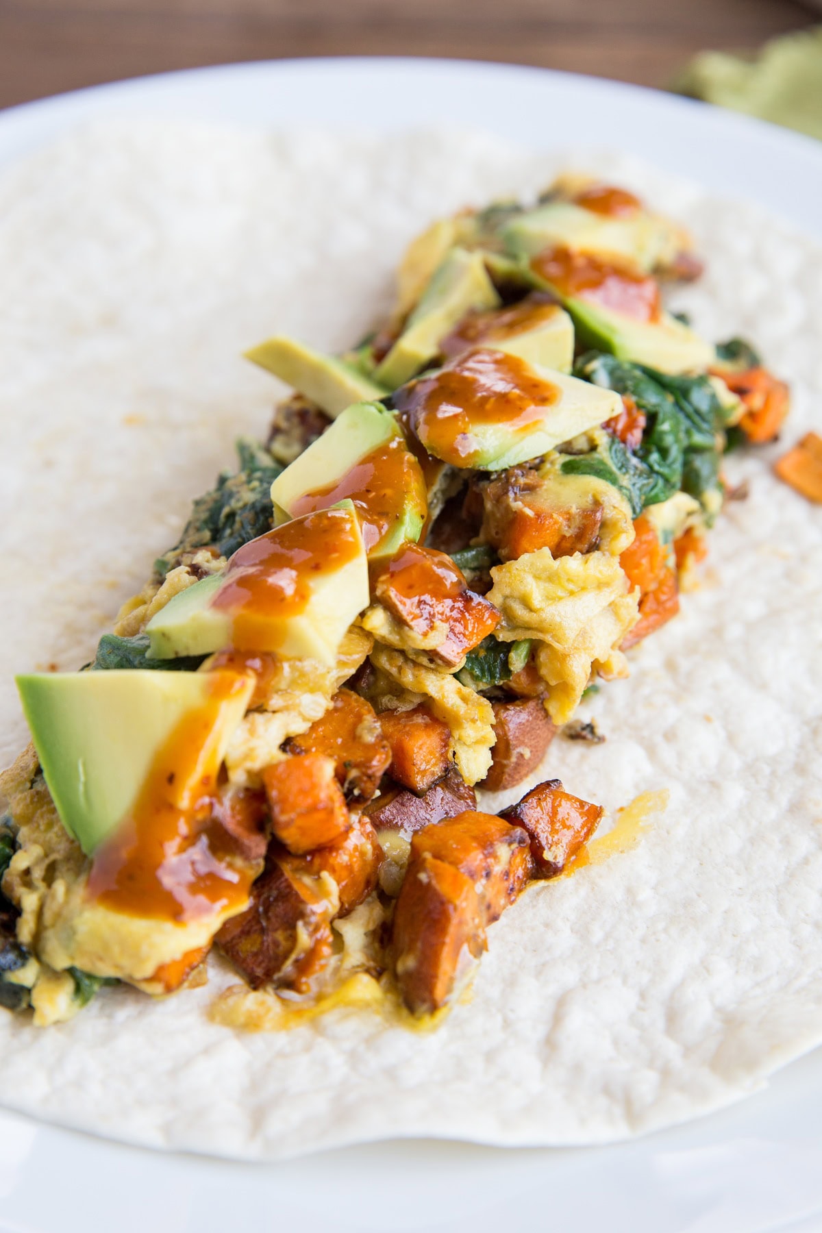 Flour tortilla on a plate with scrambled eggs, sweet potato, spinach, and zucchini in the middle with diced avocado and hot sauce on top.