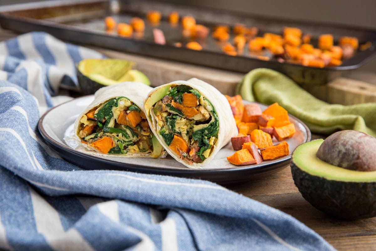 Vegetable breakfast burrito on a plate cut in half so you can see the inside. Roasted sweet potatoes to the side on the plate and half of an avocado to the side with a blue striped napkin.