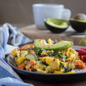 Plate of roasted vegetable scramble steaming with a mug of coffee or tea in the background. Sliced avocado on top of the scramble and sliced strawberries on the side of the plate.