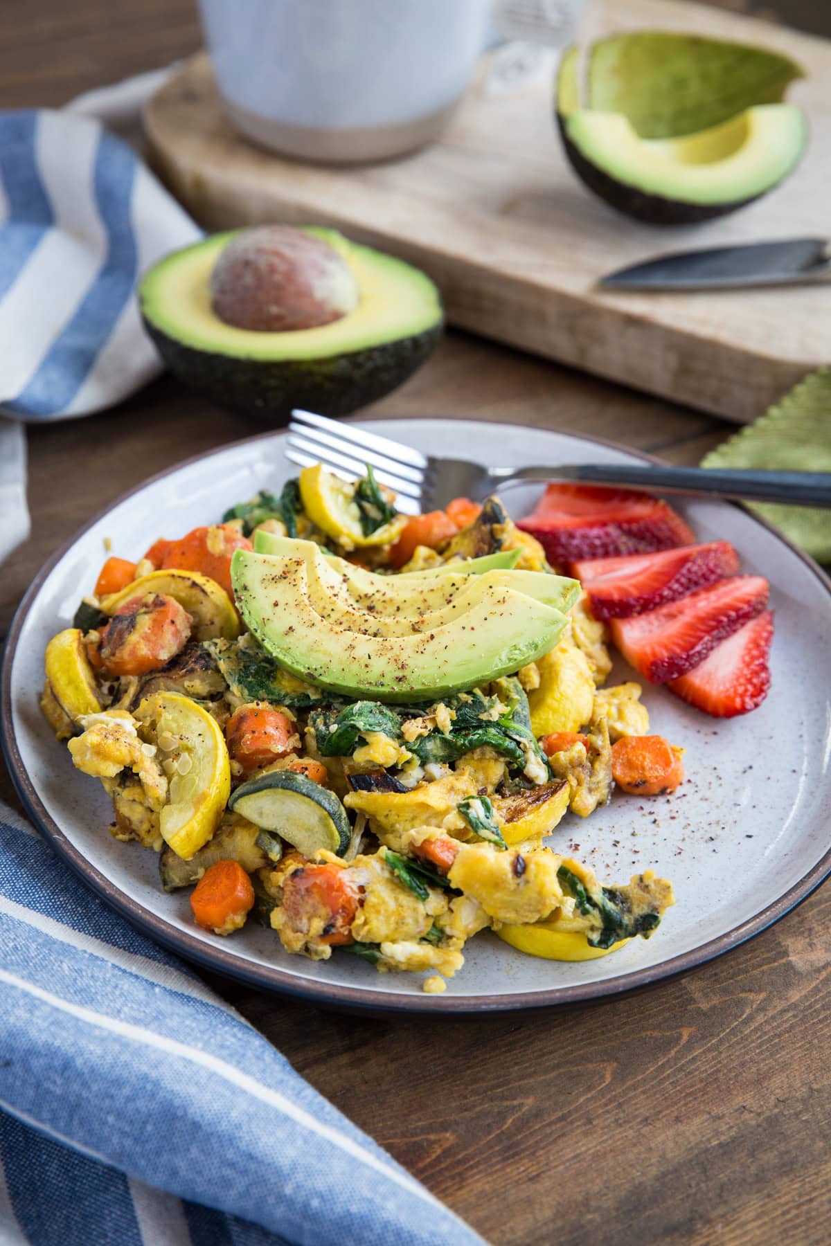 Plate of scrambled eggs with roasted vegetables cooked inside with sliced avocado on top and sliced strawberries to the side. Ready to eat for breakfast.