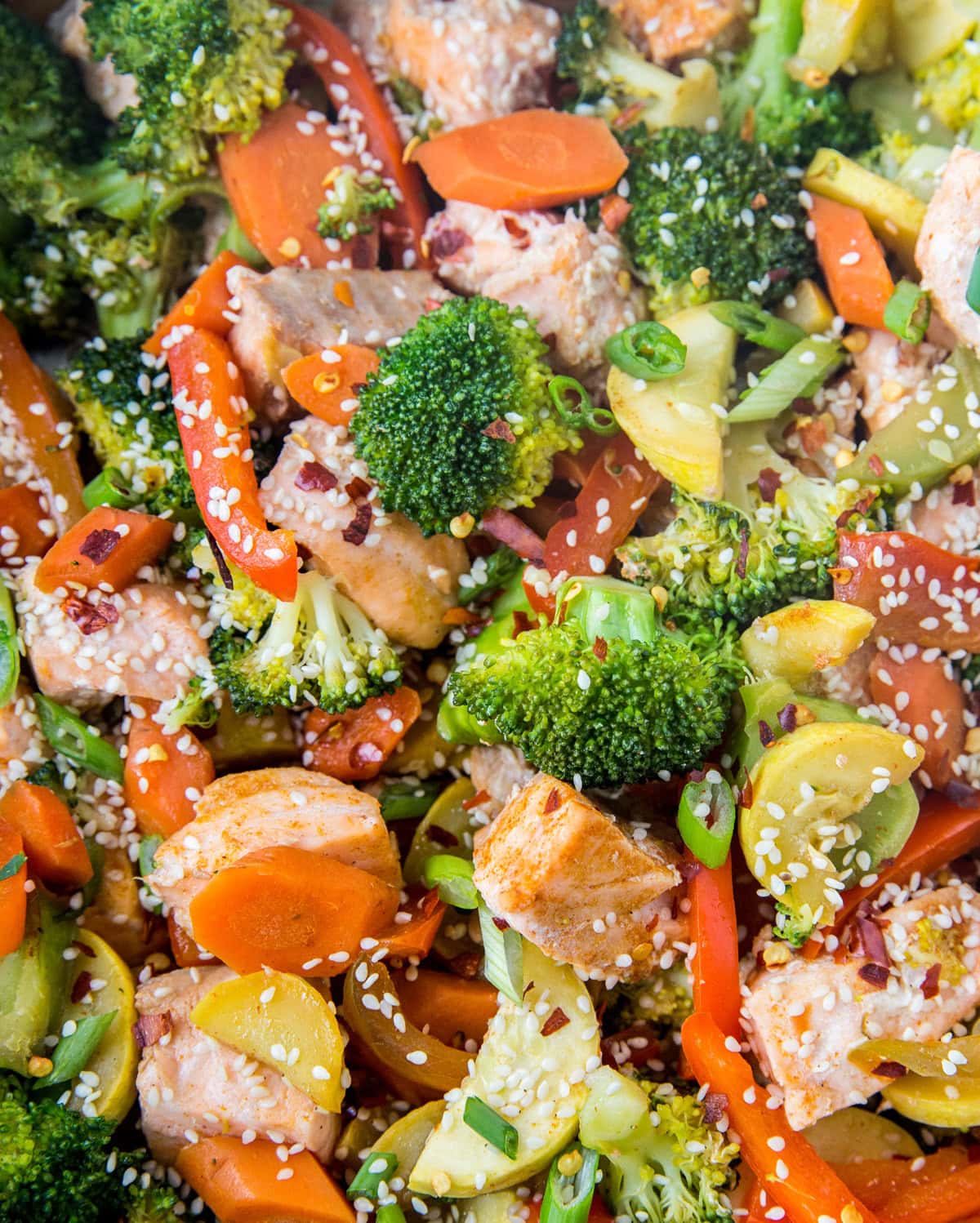 Closeup image of salmon stir fry in a skillet, sprinkled with sesame seeds, green onions, and red pepper flakes.