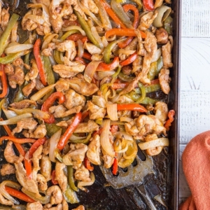Sheet pan with chicken fajitas on top with a metal spatula scooping some off and a red napkin to the side.