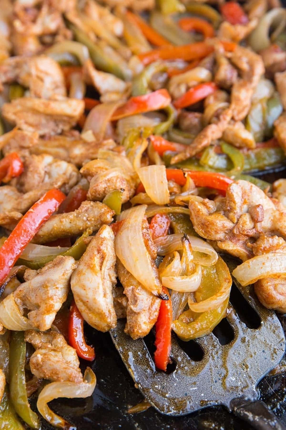baking sheet full of chicken, onions, bell peppers and seasonings with a metal spatula scooping some off the sheet.
