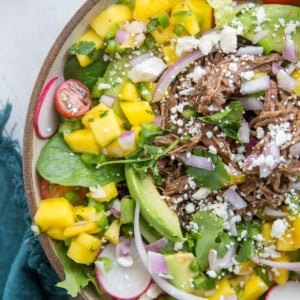 Shredded Beef Taco Salad with mango salsa, queso fresco, avocado and cilantro-lime dressing in a bowl with fresh veggies and a blue napkin to the side.
