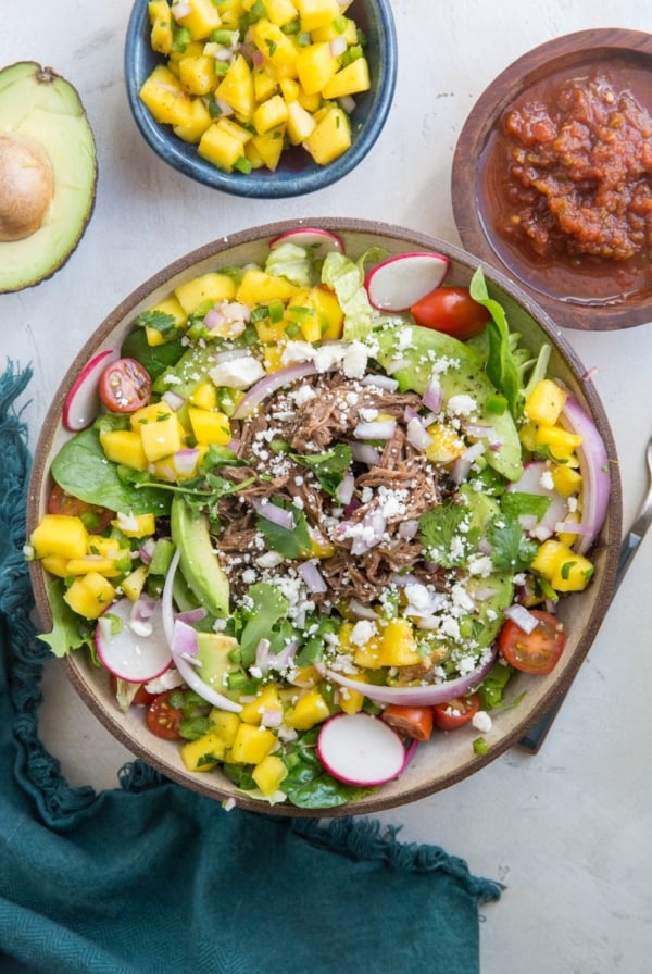 Large bowl of Shredded Beef Taco Salad with spring greens, avocado, cherry tomatoes, mango salsa, and more. A blue napkin to the side and a bowl of salsa.