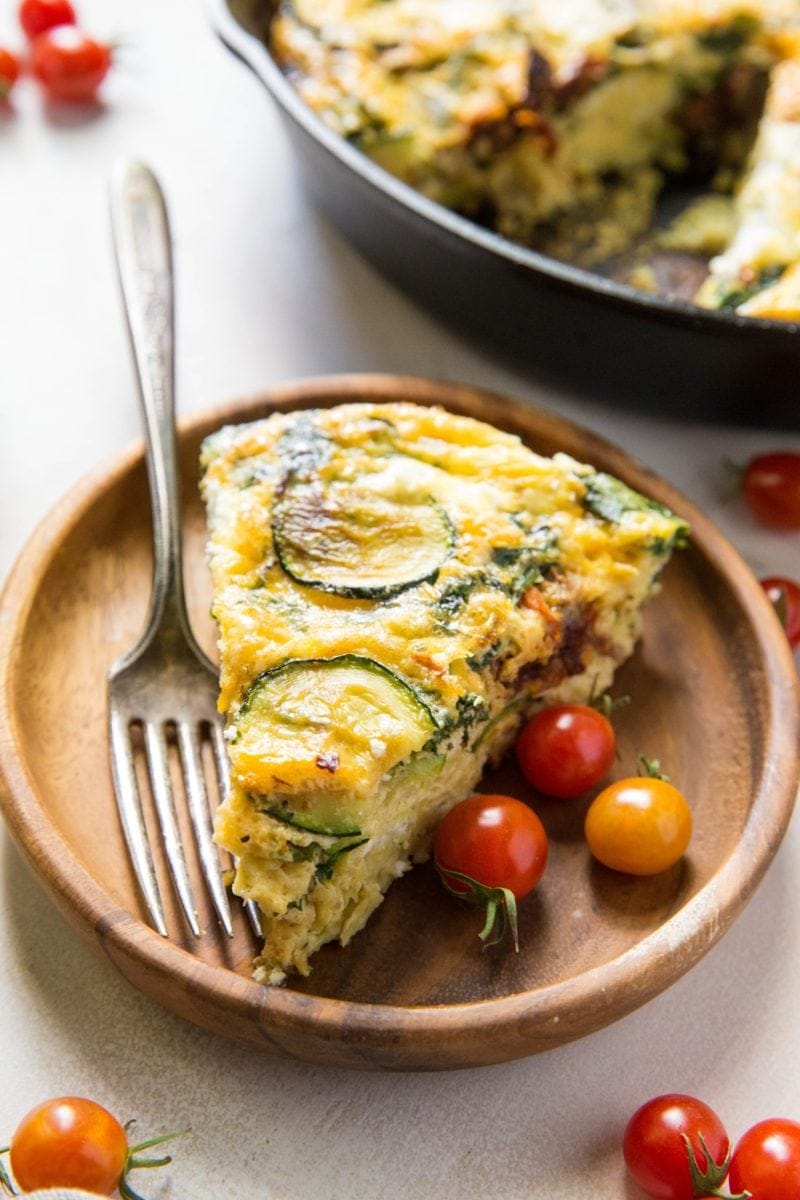 Slice of Spinach Frittata with Zucchini, Sun-Dried Tomatoes, and Goat Cheese on a plate with the rest of the frittata in the background.