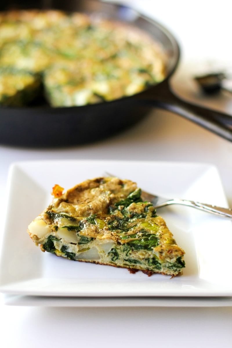 Slice of frittata on a white square plate with the rest of the frittata in a cast iron skillet in the background.