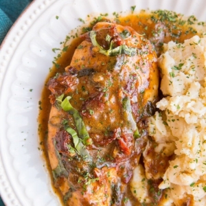 Baked Creamy Sun-Dried Tomato Chicken on a white plate with a side of mashed potatoes. A blue napkin to the side and fresh basil.